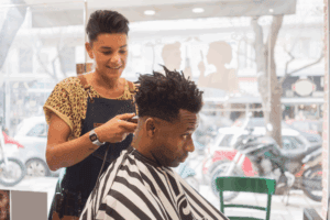 A young woman smiling while cutting the hair of a male client in a barber shop.